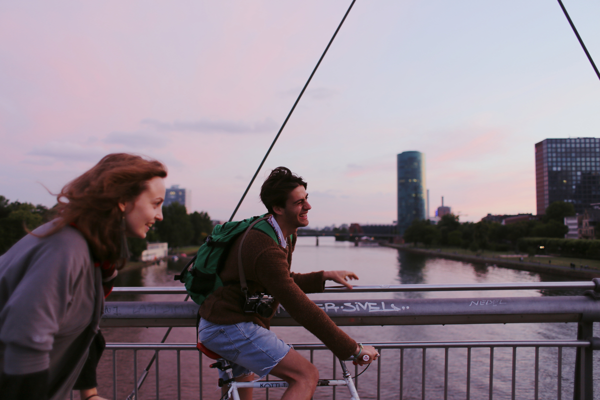 A couple biking on a bridge over a river in the early morning with tall buildings visible on the shoreline.